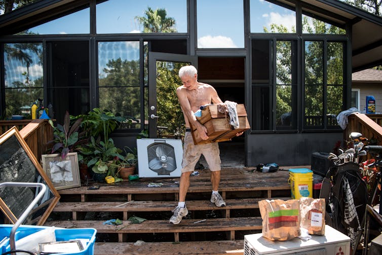 A man carries boxes out of a house that flooded up to its second story.